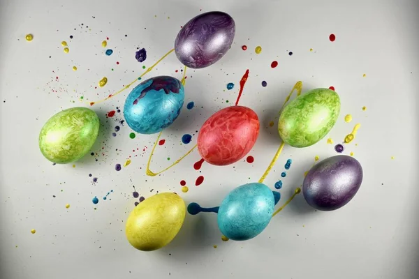 Multi-colored painted eggs on a light surface filled with drops of multi-colored paint. View from above. — Stock Photo, Image