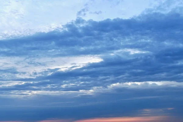 Cielo azul suave con una delgada franja rosada de puesta de sol en la esquina inferior derecha. Nubes azules brillantes estiradas horizontalmente y ligeramente diagonalmente hacia arriba a la derecha, dejando aberturas celestes brillantes . — Foto de Stock