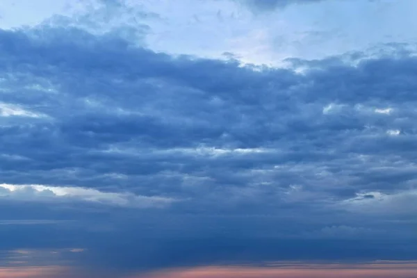 Nubes azules alargadas horizontalmente contra el cielo azul. Arriba está la parte libre del firmamento. Debajo hay una franja de luz de melocotón rosado, similar al rastro de un sol pasado . — Foto de Stock