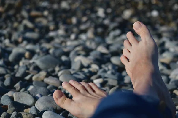 Two legs with bare feet, an extended cross on a cross, on a pebble beach. Illuminated by sunlight. In the bottom right corner of the frame.