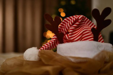 Yeni yıl kırmızı çizgili, tüylü ve ponponu olan, dikey geyik boynuzlarıyla süslenmiş, Noel ağacı çelenklerinin sıcak ışıklarına sarılı, kırmızı şeritli bir şapka..