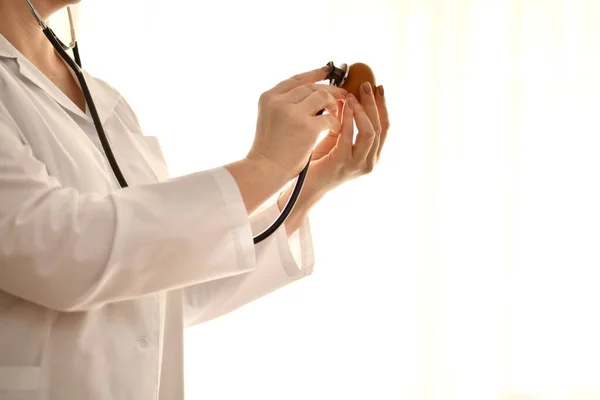 Doktor úr, egy nő fehér köpenyben sztetoszkóppal figyelmesen hallgatja egy tyúktojás szívverését a kagylón keresztül.. — Stock Fotó