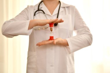 Doktor, beyaz cüppeli bir kadın, önünde duruyor ve iki elin avuçlarının arasında parlak bir arka planda, bir kum saati tutuyor.. 