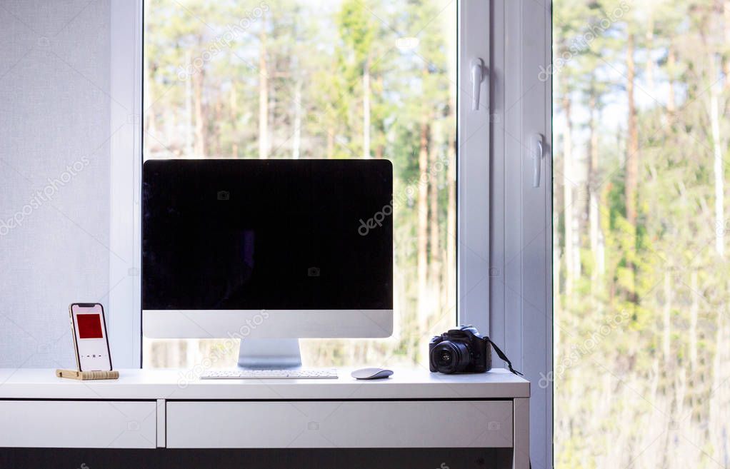 Desktop of a photographer: computer, camera, phone, mouse and keyboard, window with summer forest outside, minimalism, the room of freelancer, minimalist flat, apartment