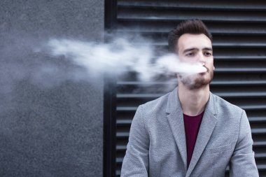 Genç dalgın adam ötüyor, duman üflüyor. Yakışıklı hipster iş adamı ceketbreak sahip, sigara, açık havada oturan. Elektronik sigara içmek, nikotin bağımlılığı. Sağlıksız yaşam tarzı, habbit.