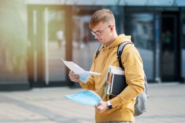 Yakışıklı bir genç, ciddi odaklanmış gözlüklü bir öğrenci kitap ve ders kitaplarıyla dışarıda duruyor, gazete okuyor, belge okuyor, metin sonuçları, yoğun bakışla muayene oluyor.