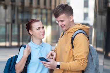 Young woman dont understand humor of her boy. Girl looking at guy with disgust, indignation. Stupid dumb humor, vulgar joke. Students with backpack looking at screen of cell mobile phone, smartphone
