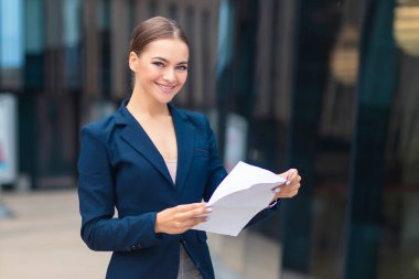 Elinde belgelerle mutlu ve başarılı bir kadın, elinde kağıtlar, takım elbiseli neşeli bir iş kadını kameraya bakarak gülümsüyor. Açık havada genç ve güzel bir iş kadını. Banka yöneticisi