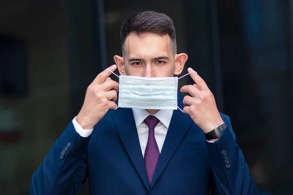 Serious businessman, young guy in suit put on protective medical mask to his face outdoors, near business office, looking at camera. Coronavirus, business, virus, pandemic covid-19 concept.