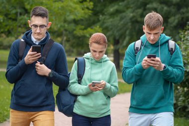 Üç öğrenci, genç milenyum çocuğu, iki erkek ve kadın, bir grup arkadaş parkta yürüyor ve cep telefonlarına bakıyorlar. Sosyal medya, internet ve akıllı telefon bağımlılığı. Modern problem