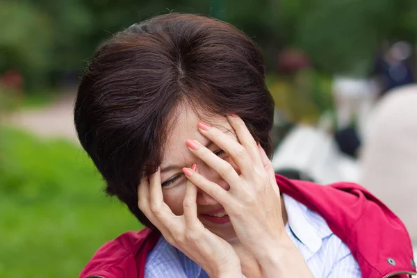 Beautiful positive emotional woman is smiling, laughing outdoors in a park, lady is covering her face with hands because of funny joke. — Stock Photo, Image