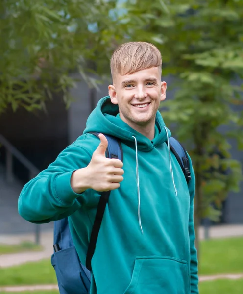 Hermosa estudiante universitaria alegre con mochila mostrando el pulgar hacia arriba, como, gesto de aprobación al aire libre en el campus. Joven feliz hombre exitoso positivo mirar a la cámara y sonreír, riendo. Entra a la universidad — Foto de Stock