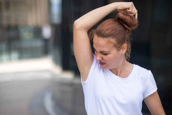 Giovane donna sudata annusare le ascelle nel disgusto in bianco T-shirt, stropicciandosi il viso a causa del cattivo odore. — Foto Stock