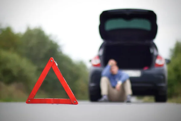 Accidente en la carretera. Triángulo de advertencia de emergencia y un hombre borroso sentado cerca del coche dañado con el tronco abierto —  Fotos de Stock