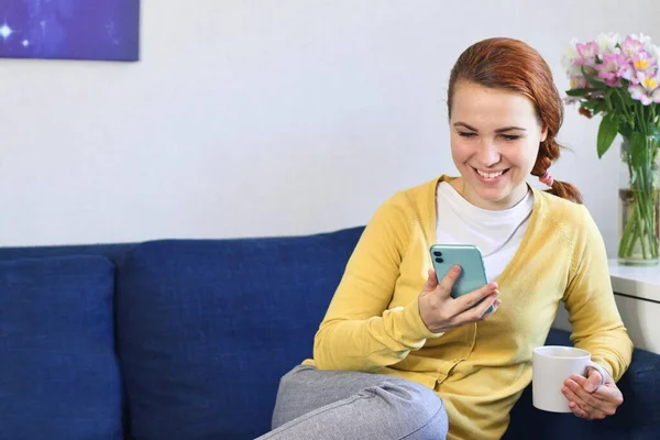 Ευτυχισμένη θετική νεαρή γυναίκα κάθεται στον καναπέ στο σαλόνι στο σπίτι, χαμογελώντας με κινητό τηλέφωνο στο χέρι, πίνοντας από ένα φλιτζάνι, κουβεντιάζοντας στα μέσα κοινωνικής δικτύωσης — Φωτογραφία Αρχείου