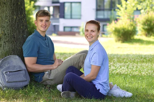 Un cuplu tânăr fericit de bărbat frumos și o femeie frumoasă stând pe o iarbă într-un parc de vară, sprijinindu-se pe un copac cu rucsac și zâmbind, uitându-se la cameră — Fotografie, imagine de stoc