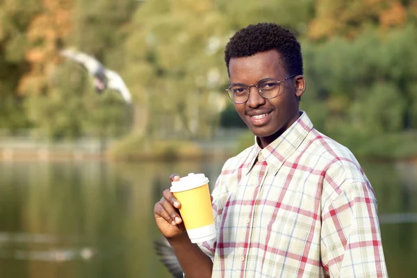 Retrato de un joven afro-americano negro feliz y positivo con camisa sonriendo en el parque de verano u otoño y bebiendo té caliente o café de una taza de plástico. Café para llevar concepto — Foto de Stock