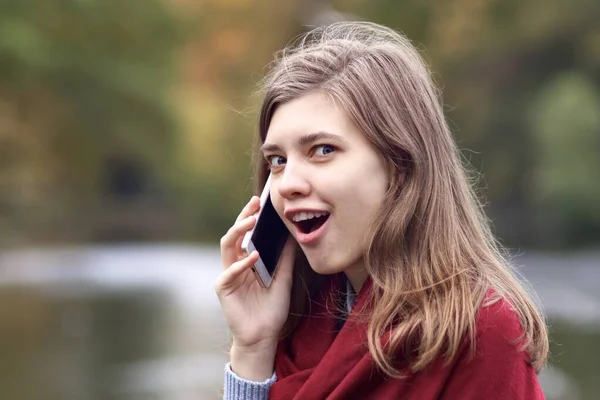 Surprised shocked happy young woman talking on cell mobile smart phone outdoors in autumn park with open mouth and excited look — Stock Photo, Image