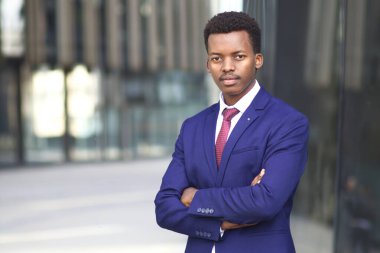Ciddi derecede kendinden emin, başarılı, Afro-Amerikalı, takım elbiseli genç bir iş adamının portresi açık hava iş yerinde ellerini çapraz yapıp kameraya bakıyor.