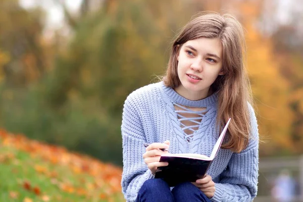 Young beautiful woman writer, teenager student girl is studying outdoors in golden autumn park writing in notebook or diary with concentrated look, doing homework