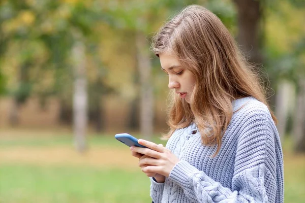Jovem bela mulher concentrada, adolescente menina está olhando para a tela de seu telefone celular, lendo ou digitando uma mensagem ao ar livre em um parque. Mídia social, vício em smartphones. — Fotografia de Stock