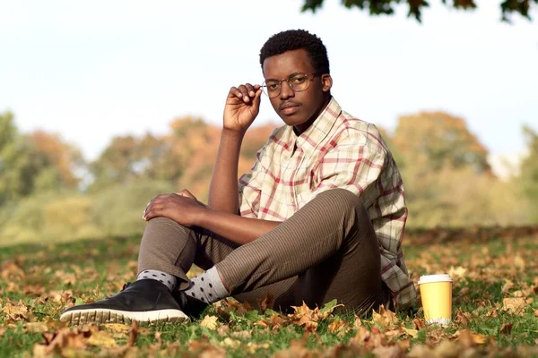 Portrait of serious intelligent handsome young black African Afro American man in glasses sitting on the grass in golden autumn park in glasses with cup of coffee, looking at camera — Stock Photo, Image