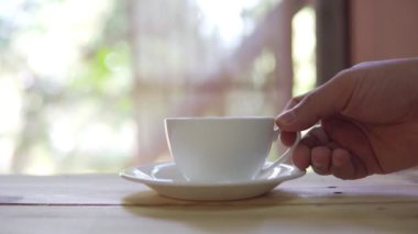 Footage B roll of hand placing hot ceramic white hot coffee cup on saucer over wooden table in nature green background. Breakfast and good food.