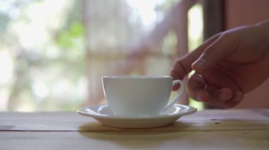 Footage B roll of hand placing hot ceramic white hot coffee cup on saucer over wooden table in nature green background. Breakfast and good food.