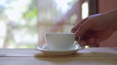 Footage B roll of hand placing hot ceramic white hot coffee cup on saucer over wooden table in nature green background. Breakfast and good food.