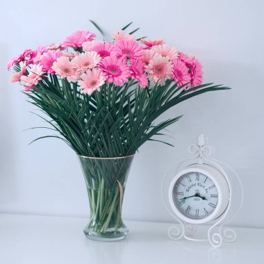 Yakın plan beyaz alarm saati ve masanın üzerindeki cam vazoda çiçek buketi. Beyaz arka plan. Durgun hayat.