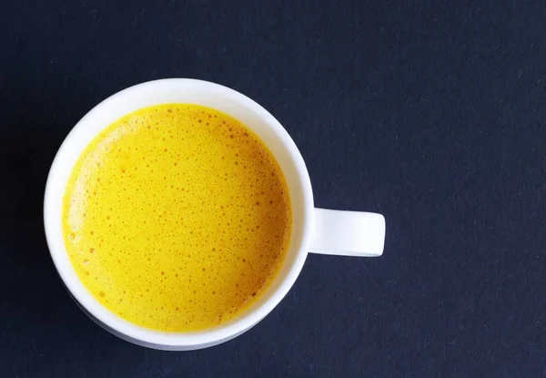 Golden (turmeric) milk in a white cup on the dark background. Top view.