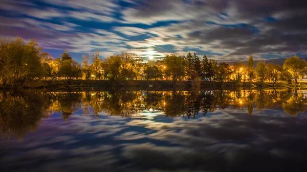 Şehir ışıkları ve bulutlar tarafından aydınlatılan ağaçlarla nehrin üzerindeki gece manzarası