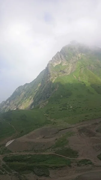 Na encosta de uma montanha alta com uma encosta verde e um topo em nuvens e nevoeiro. Esportes e base turística nas montanhas. Krasnaya Polyana, Sochi, Rússia . — Fotografia de Stock