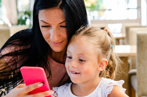 Gülümseyen bir kız akıllı telefondan tatlı bir kızla selfie çekiyor, mutlu bir genç anne gülüyor, küçük kızıyla fotoğraf çekiyor, telefonla oynuyor, cep telefonuyla aile videosu kaydediyor.