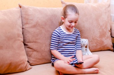 Evdeki kanepede oturan ve elinde cep telefonu tutan tatlı bir kız. Cep telefonundaki uzaktan iletişim, aile videolarını kaydediyor. Evde uzaklık öğrenimi.