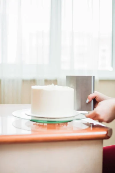 Confectioner using a knife to smooth the sides and top of the cake, close-up side view photo. The confectioner smoothes the white cream on the biscuit cake using a culinary spatula. The concept of home baking, cooking cakes. process of decorating the