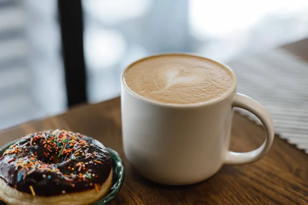 Coffee with a drawn heart and milk on a wooden table in a coffee shop. Pink donut with scattering on the table next to the coffee