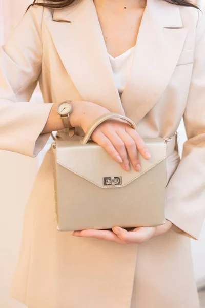 bag in hand. Vertical photo of a girl with a gold bag in her hands and watches. Beige suit