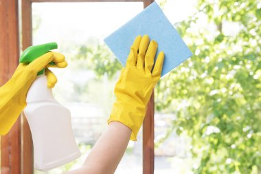Kız ev hanımı sarı lastik eldiven ve mavi bir bez ile evin pencereleri yıkar. Temizlik ve ev temizliği. Arka plan penceresi - yeşil yapraklar. Ahşap ve plastik pencereler