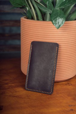 Ahşap arka plan üzerinde maço kahverengi deri telefon kılıfı. Elle yapılmış. Saksı çiçeğinin arka planına karşı el yapımı telefon kılıfı