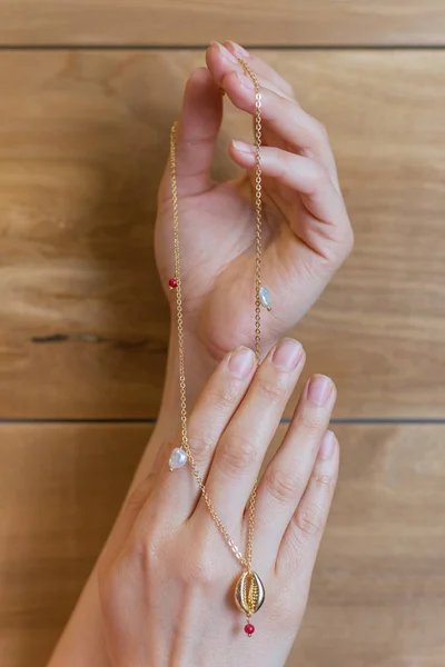 Expensive beautiful jewelry with pearls and a seashell of gold in the hands of a girl with a natural manicure on a wooden background. Luxury pendant on the neck in the hands of the girl.