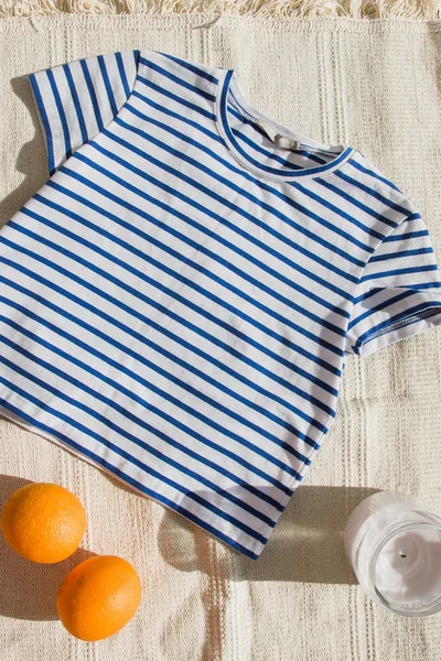 Fashionable cropped top with blue stripes lies on the light carpet. Shirt next to an orange and a candle. Stylish vest lies on a light background