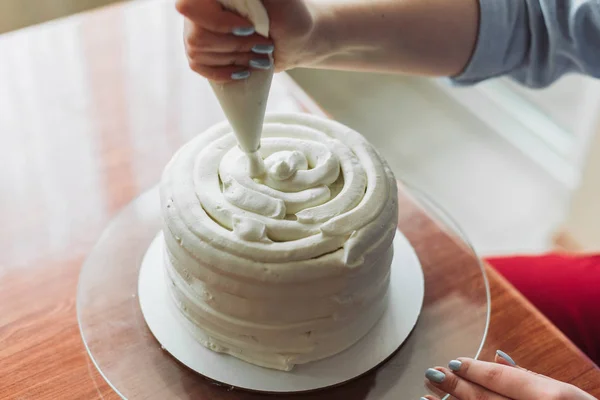 A lovely girl making a cake in a bakery. The girl smoothes the cream on the cake. White cake on a wooden table.