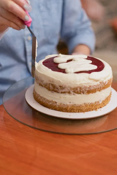 Krásná holka, co dělá dort v pekárně. pekař na červenou koláč tlačí smetanu. Bílý dort na dřevěném stole. — Stock fotografie