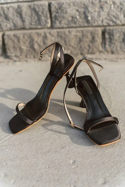 Black leather open sandals with heels stand on the asphalt in the sun. Women's shoes. Vertical photo of stylish and fashionable high heel shoes. — Stock Photo, Image
