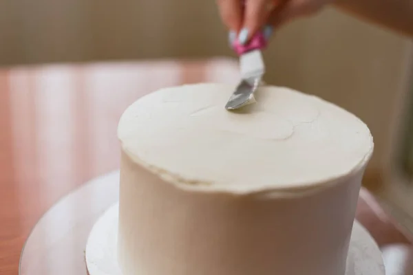 A rapariga aperta o creme no bolo. Uma rapariga adorável a fazer um bolo numa padaria. A menina suaviza o creme no bolo. Bolo branco em uma mesa de madeira — Fotografia de Stock