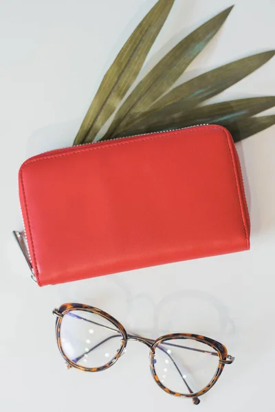 Das rote schicke Portemonnaie und die Modebrille der Frau liegen mit einem Palmblatt auf dem Tisch. das Konzept von Stil, Mode und weiblichem Selbstvertrauen — Stockfoto