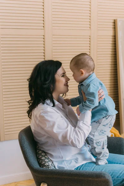 Eine junge Mutter in Jeans und weißem Hemd mit einem Baby auf dem Arm amüsiert sich und spielt. Die Liebe der Mutter — Stockfoto