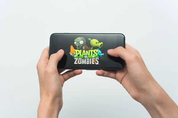 Samara, Russia -07, 29, 2019: Teenage boy holding a phone in his hands with a game plants vs zombies mobile on a white background. A young guy plays a plants vs. zombies game on Iphone 8 Plus — Stock Photo, Image