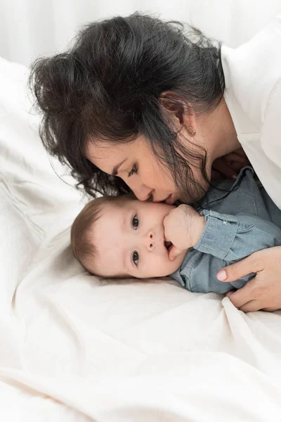 Die dunkelhaarige Mutter küsst ein einjähriges Baby in einem hellen Bett. Mamas Liebe zu ihrem Sohn — Stockfoto